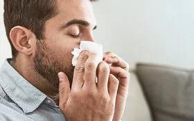 Ein Mann schnäubt sich die Nase wegen einer Hausstaubmilbenallergie.