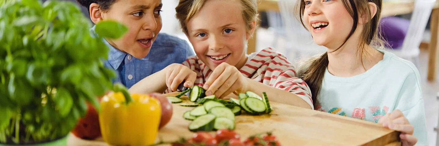 Drei lachende Kinder schneiden Gemüse.