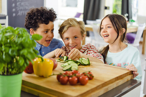 Drei lachende Kinder schneiden Gemüse.