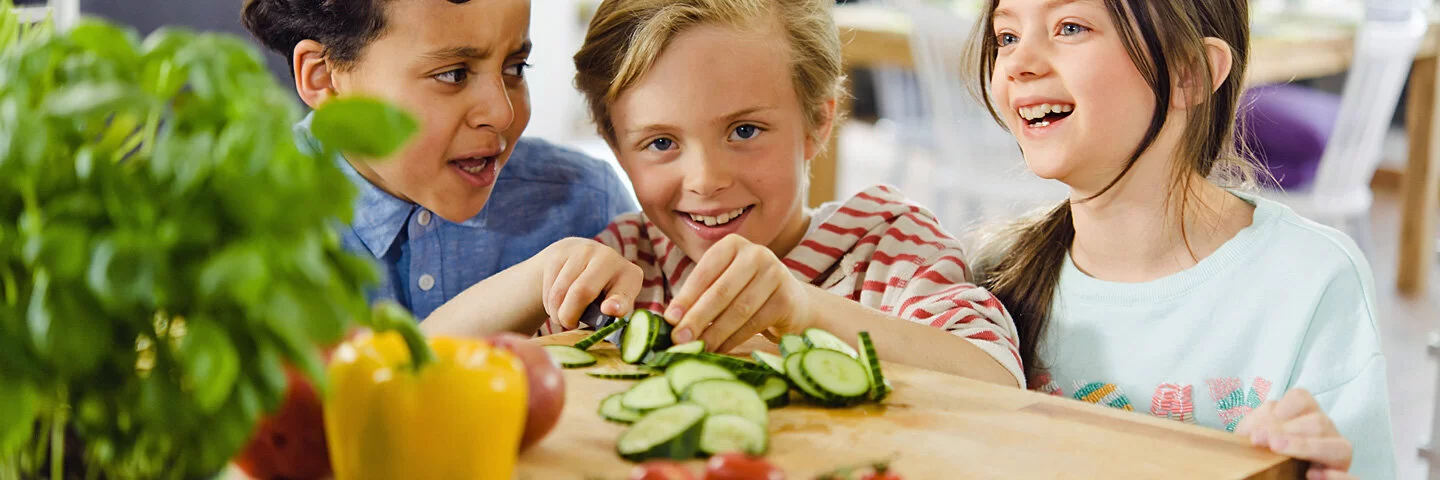Drei lachende Kinder schneiden Gemüse.
