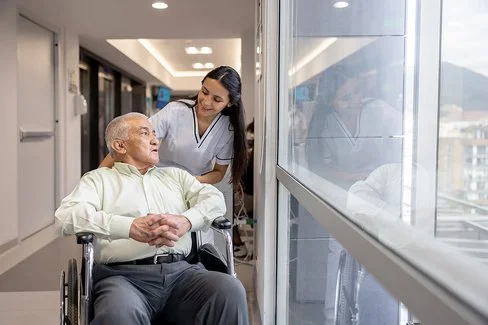 Eine weibliche Pflegefachperson fährt einen Patienten im Rollstuhl durch den Flur.