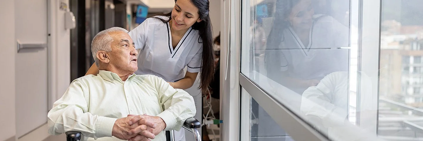 Eine weibliche Pflegefachperson fährt einen Patienten im Rollstuhl durch den Flur.