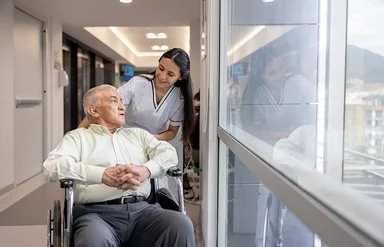 Eine weibliche Pflegefachperson fährt einen Patienten im Rollstuhl durch den Flur.