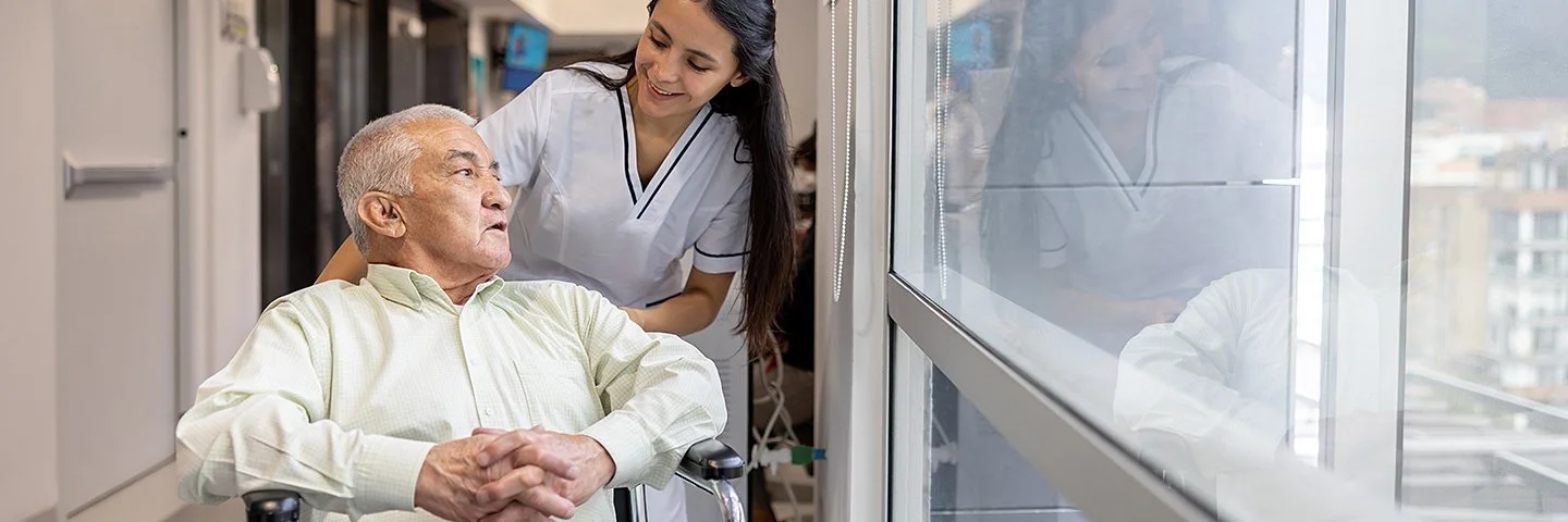 Eine weibliche Pflegefachperson fährt einen Patienten im Rollstuhl durch den Flur.