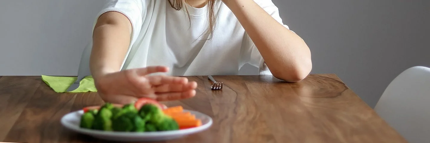 Junges Mädchen sitzt erschöpft am Tisch und schiebt einen Teller mit Gemüse von sich weg – sichtbar ohne Appetit.