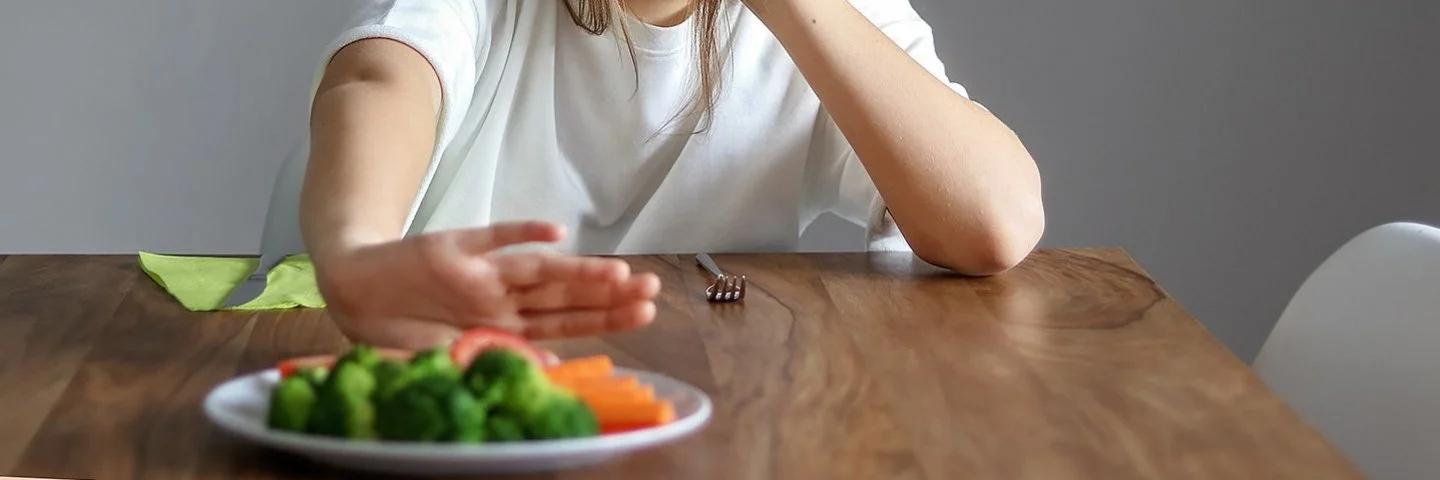 Junges Mädchen sitzt erschöpft am Tisch und schiebt einen Teller mit Gemüse von sich weg – sichtbar ohne Appetit.