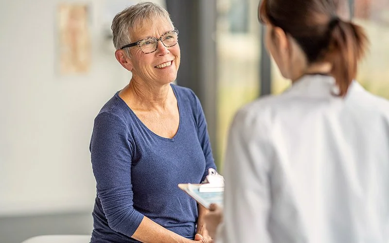 Eine Ärztin beträt eine Seniorin in der Sprechstunde. Die Seniorin lächelt die Ärztin an, die Ärztin ist von hinten zu sehen.