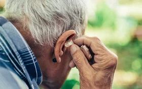 Ein älterer Mann sitzt im Garten und steckt sein Hörgerät ins Ohr.