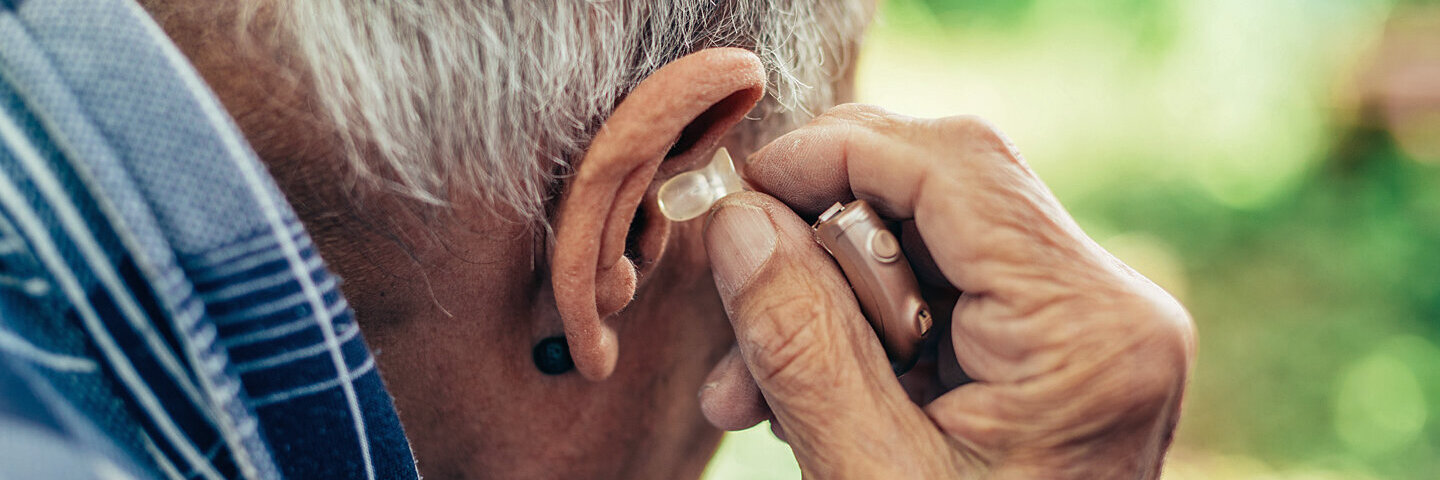 Ein älterer Mann sitzt im Garten und steckt sein Hörgerät ins Ohr.
