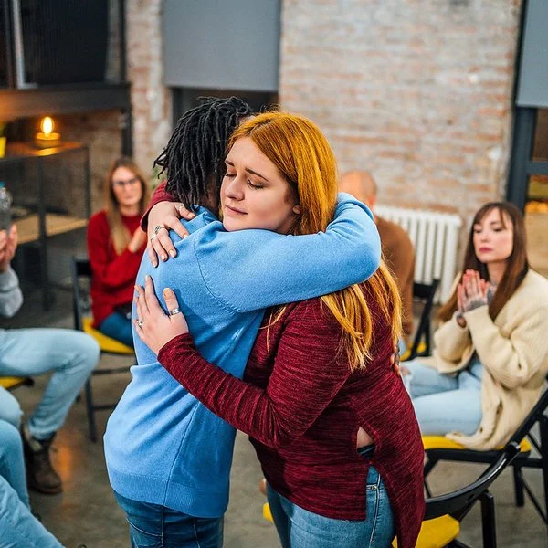 Frauen und Männer sitzen in einem Stuhlkreis zusammen. Eine junge Frau umarmt eine andere und begrüßt sie als neues Mitglied.