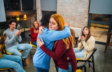 Frauen und Männer sitzen in einem Stuhlkreis zusammen. Eine junge Frau umarmt eine andere und begrüßt sie als neues Mitglied.
