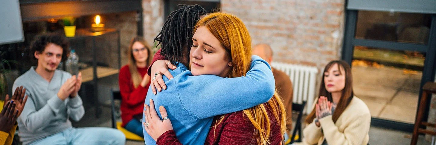 Frauen und Männer sitzen in einem Stuhlkreis zusammen. Eine junge Frau umarmt eine andere und begrüßt sie als neues Mitglied.