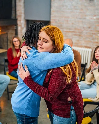 Frauen und Männer sitzen in einem Stuhlkreis zusammen. Eine junge Frau umarmt eine andere und begrüßt sie als neues Mitglied.