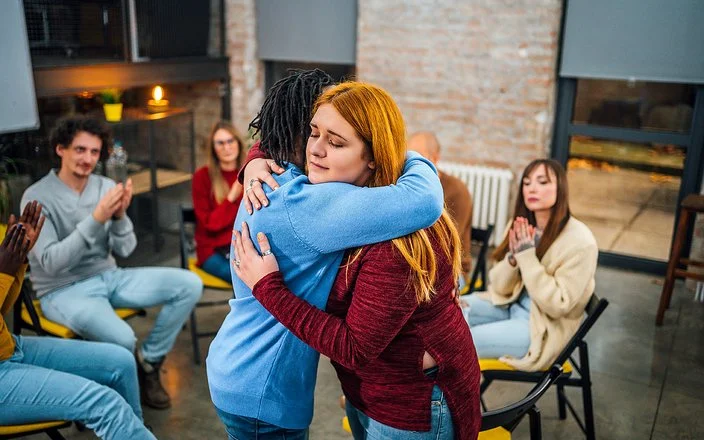 Frauen und Männer sitzen in einem Stuhlkreis zusammen. Eine junge Frau umarmt eine andere und begrüßt sie als neues Mitglied.