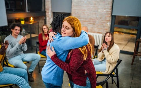Frauen und Männer sitzen in einem Stuhlkreis zusammen. Eine junge Frau umarmt eine andere und begrüßt sie als neues Mitglied.