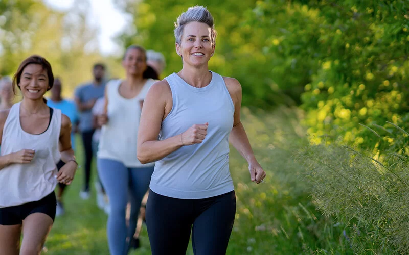 Mehrere junge bis mittelalte Frauen, die sportliche Kleidung tragen und in einer ländlichen Umgebung joggen. 