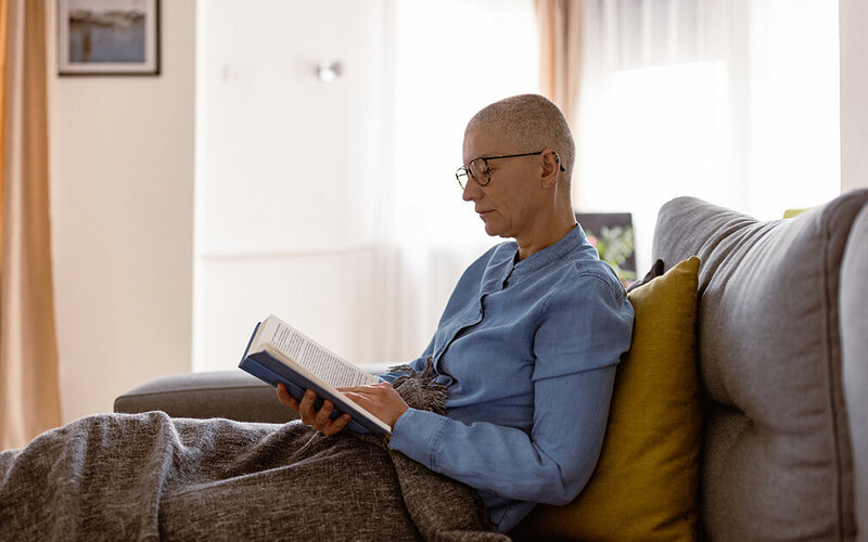 Eine Krebspatientin mit kurzen Haaren sitzt in eine Decke gehüllt auf der Couch und liest ein Buch.