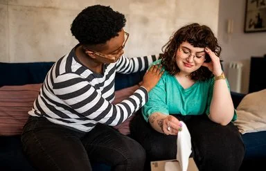 Zwei Frauen sitzen auf einem Sofa, eine davon legt der anderen tröstend die Hand auf die Schulter – die andere Frau schaut traurig.