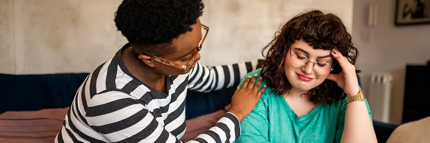 Zwei Frauen sitzen auf einem Sofa, eine davon legt der anderen tröstend die Hand auf die Schulter – die andere Frau schaut traurig.