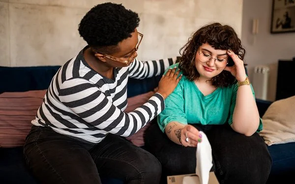 Zwei Frauen sitzen auf einem Sofa, eine davon legt der anderen tröstend die Hand auf die Schulter – die andere Frau schaut traurig.