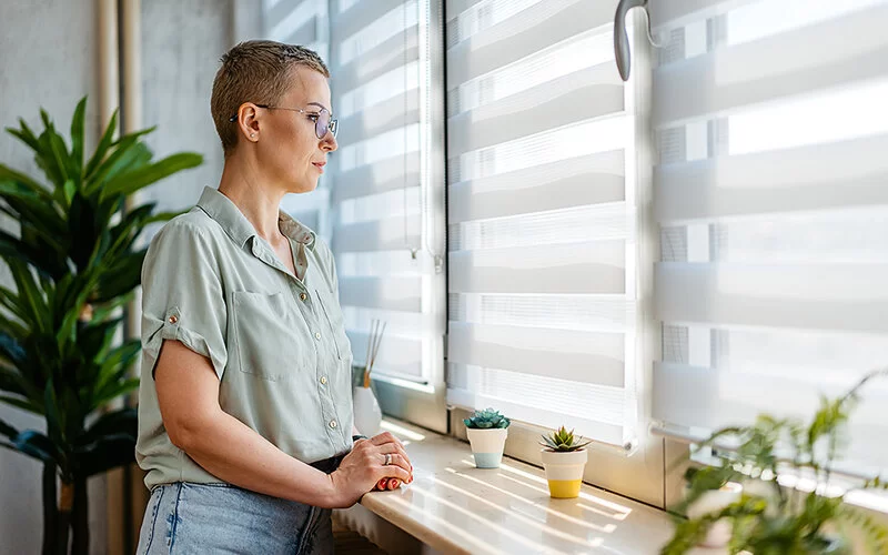 Frau mit kurzen Haaren und Brille steht vor einem Fenster mit Rollos und schaut heraus.
