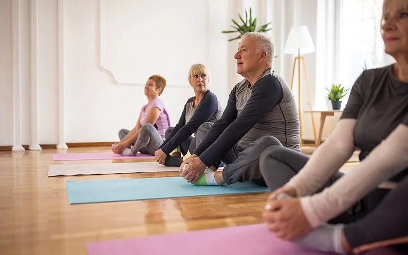 Drei Frauen und ein Mann in mittlerem Alter sitzen in sportlicher Kleidung auf Matten und praktizieren Yoga.