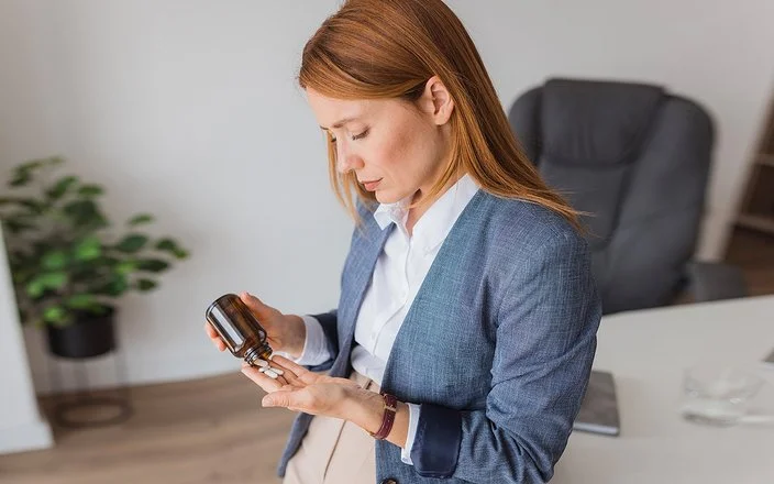 Eine Frau steht vor ihrem Schreibtisch im Büro. Sie schüttet aus einem Tablettenglas Tabletten in ihre Handfläche.