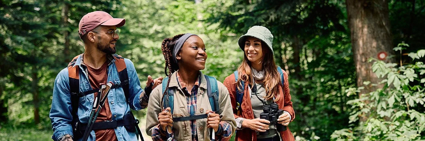 Zwei Frauen und ein Mann wandern mit Rucksäcken und Trekkingstöcken auf einem grünen Waldweg.