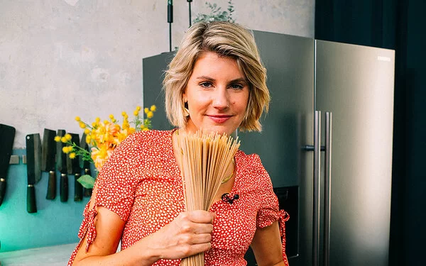 TV-Köchin Felicitas Then hält eine Handvoll Vollkorn-Spaghetti vor der Brust und präsentiert diese freundlich lachend in die Kamera.