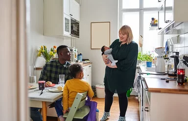 Eine Mutter mit Säugling auf dem Arm steht in der Küche, zwei weitere Personen sitzen beim Frühstück am Küchentisch. Die Ernährung in der Stillzeit ist sehr wichtig für die Entwicklung des Kindes.