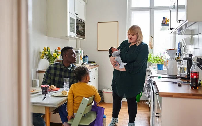 Eine Mutter mit Säugling auf dem Arm steht in der Küche, zwei weitere Personen sitzen beim Frühstück am Küchentisch. Die Ernährung in der Stillzeit ist sehr wichtig für die Entwicklung des Kindes.