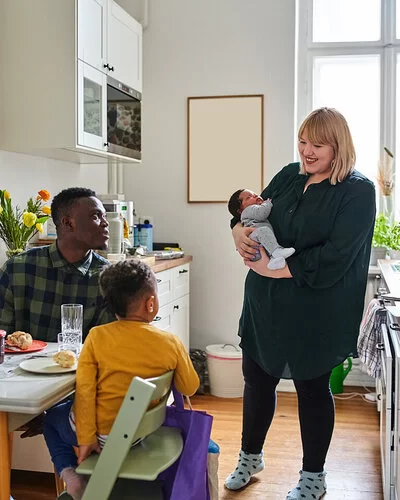 Eine Mutter mit Säugling auf dem Arm steht in der Küche, zwei weitere Personen sitzen beim Frühstück am Küchentisch. Die Ernährung in der Stillzeit ist sehr wichtig für die Entwicklung des Kindes.