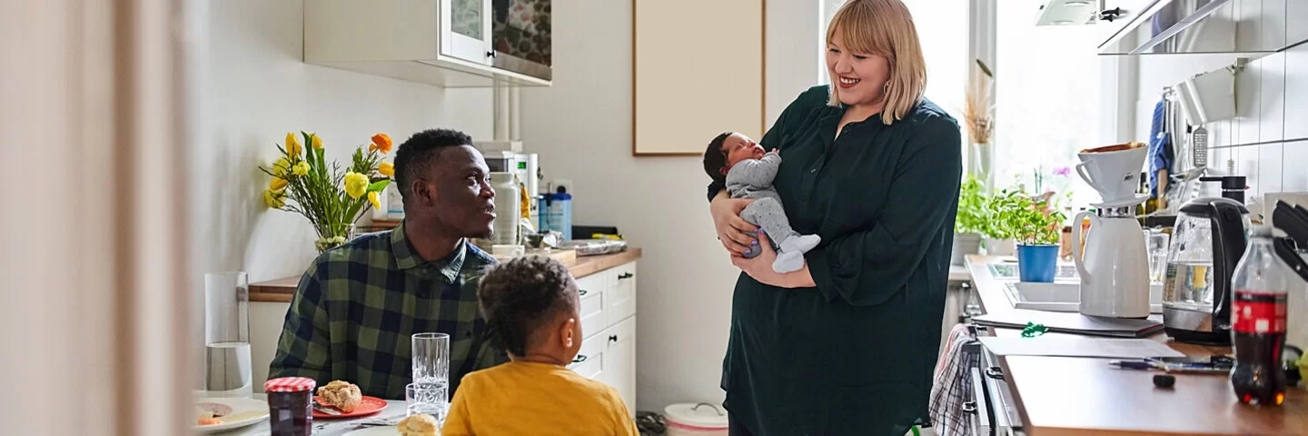 Eine Mutter mit Säugling auf dem Arm steht in der Küche, zwei weitere Personen sitzen beim Frühstück am Küchentisch. Die Ernährung in der Stillzeit ist sehr wichtig für die Entwicklung des Kindes.