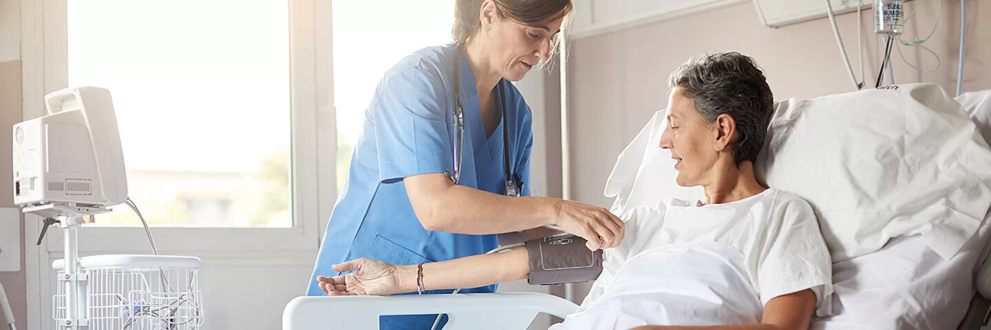 Eine Frau mittleren Alters liegt in einem Krankenbett im Krankenhaus. Eine Krankenschwester steht neben ihrem Bett und misst ihren Blutdruck am rechten Oberarm mit einem Blutdruckmessgerät.