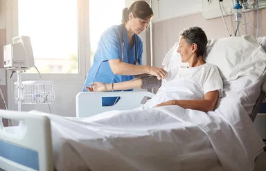 Eine Frau mittleren Alters liegt in einem Krankenbett im Krankenhaus. Eine Krankenschwester steht neben ihrem Bett und misst ihren Blutdruck am rechten Oberarm mit einem Blutdruckmessgerät.