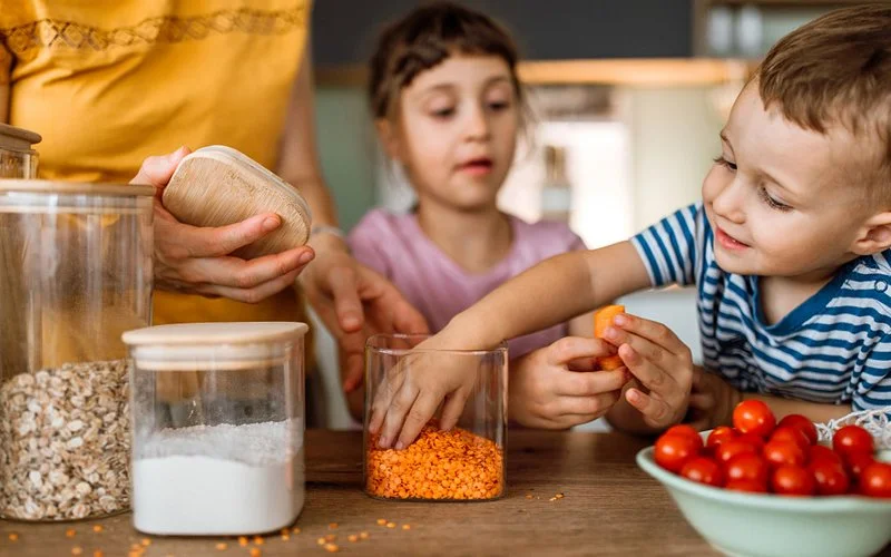 Zwei Kinder helfen beim Zubereiten einer Mahlzeit und greifen in ein Glas mit roten Linsen.