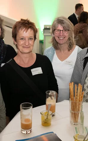 Zwei Frauen stehen an einem Stehtisch und lächeln für die Kamera.