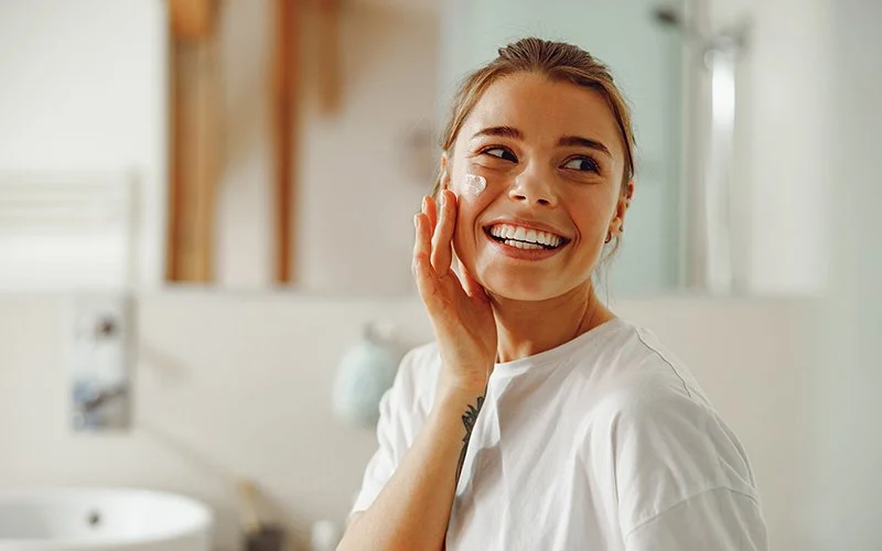 Eine junge Frau in einem Badezimmer. Sie lacht fröhlich und cremt sich die rechte Wange ein.