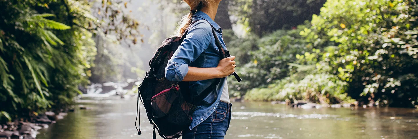 Eine Frau mit Rucksack genießt die handyfreie Zeit an einem Wasserlauf.