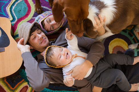 Eine junge Familie mit einem Baby liegt lachend am Boden. Ihr Familienhund beugt sich über sie.