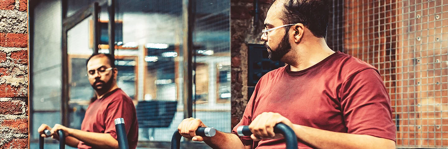Ein übergewichtiger Mann mittleren Alters betrachtet sich beim Trainieren im Fitnessstudio von der Seite im Spiegel.