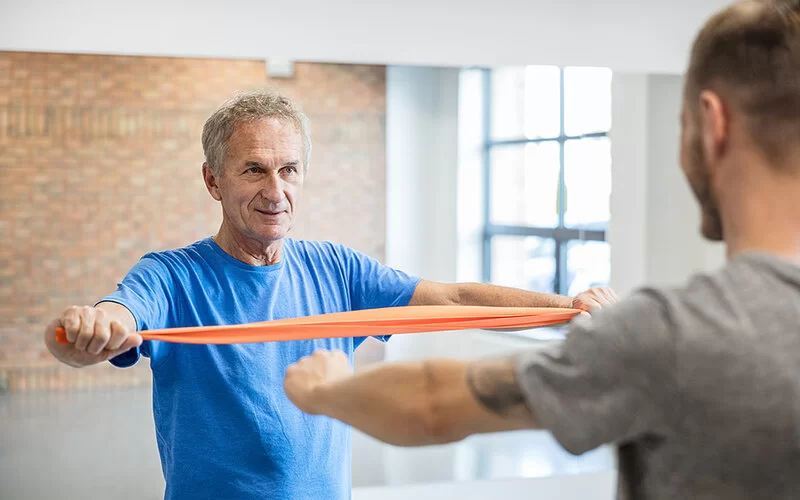 Ein Mann in einem blauen T-Shirt trainiert unter Anleitung eines Trainers mit einem orangefarbenen Widerstandsband.