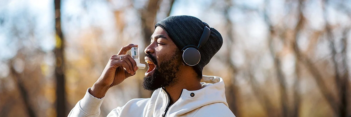 Junger Mann, der in Sportkleidung im Park steht und einen Asthma-Inhalator benutzt.