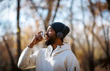 Junger Mann, der in Sportkleidung im Park steht und einen Asthma-Inhalator benutzt.