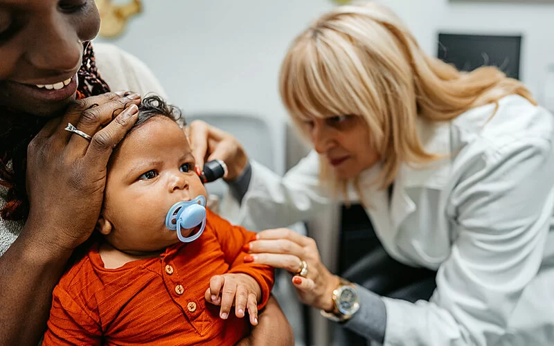 Ein Baby mit Schnuller sitzt auf dem Schoß seiner Mutter, während eine Ärztin sein linkes Ohr mit einem Otoskop untersucht.