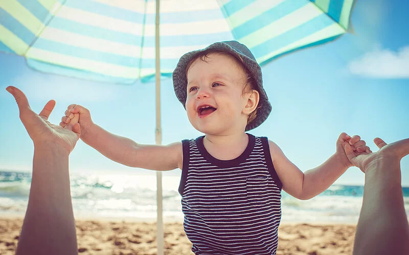 Ein lachendes Kleinkind mit Sonnenhut hält am Strand die Hände eines nicht sichtbaren Erwachsenen unter einem Sonnenschirm.