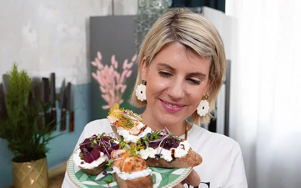 TV-Köchin Felicitas Then hält einen Teller mit vier Crostini in ihren Händen und präsentiert diese freundlich lachend in die Kamera.