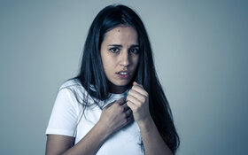 Eine junge Frau mit dunklen langen Haaren blickt erschrocken und ängstlich, die Hände hält sie schützend vor den Brustkorb.