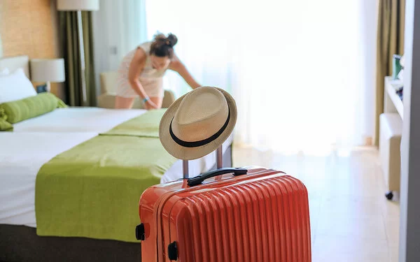 Ankunftsszene in einem Hotelzimmer: Im Vordergrund steht ein orangefarbener Rollkoffer. Auf dem Griff, den man zum Ziehen ausfahren kann, hängt ein heller Männerstrohhut. Im Hintergrund zupft eine Frau mit dunklen, zu einem Knoten zusammengefassten Haaren an der Bettwäsche eines Doppelbettes.