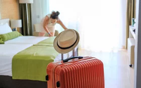 Ankunftsszene in einem Hotelzimmer: Im Vordergrund steht ein orangefarbener Rollkoffer. Auf dem Griff, den man zum Ziehen ausfahren kann, hängt ein heller Männerstrohhut. Im Hintergrund zupft eine Frau mit dunklen, zu einem Knoten zusammengefassten Haaren an der Bettwäsche eines Doppelbettes.